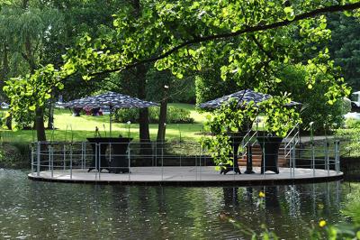 Jardin verdoyant avec arbres, pelouse soignée, tables hautes sous parasols et mobilier coloré près d'un plan d'eau.