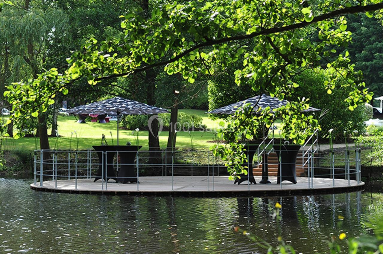 Plateforme ronde en bois sur un étang, avec parasols et mobilier, entourée de verdure.