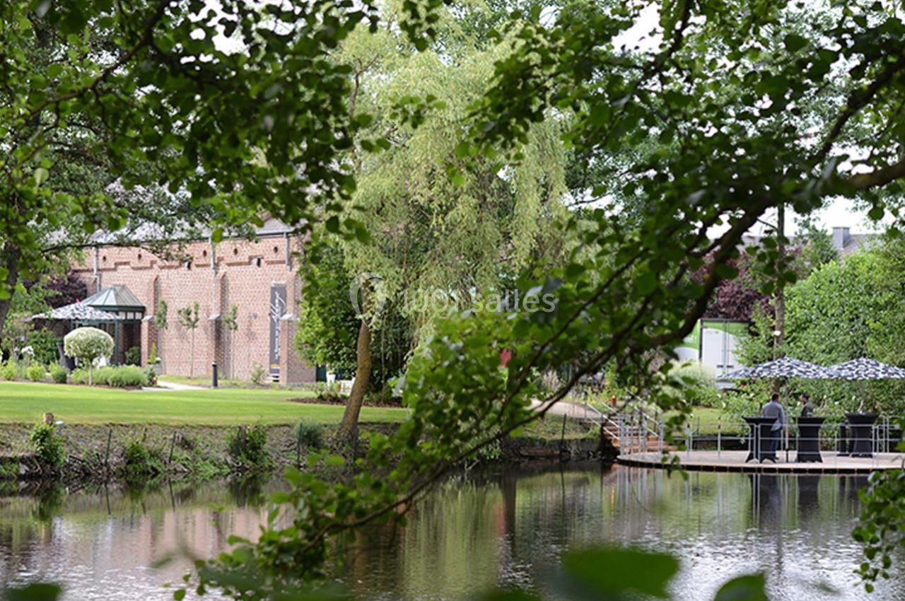 Vue d'un parc avec un étang, des arbres, un bâtiment en briques et une terrasse avec des parasols près de l'eau.