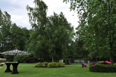 Jardin verdoyant avec arbres, pelouse soignée, tables hautes sous parasols et mobilier coloré près d'un plan d'eau.