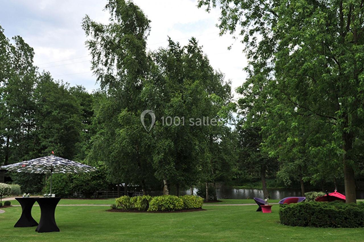 Jardin verdoyant avec arbres, pelouse soignée, tables hautes sous parasols et mobilier coloré près d'un plan d'eau.