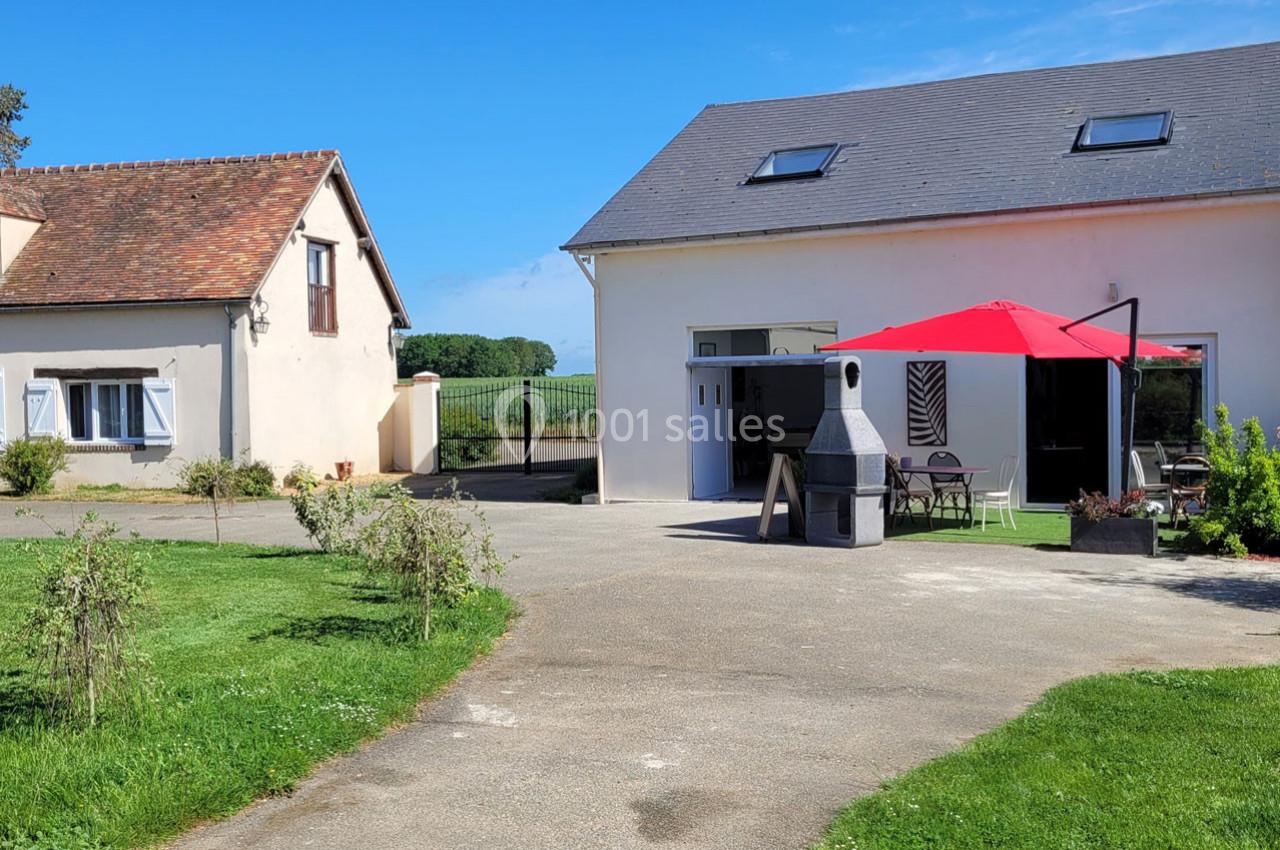 Location salle Bailleau-l'Évêque (Eure-et-Loir) - Le Domaine du Moulin 28 & SPA #10 Cour extérieure avec deux bâtiments, une table sous un parasol rouge, un barbecue et un paysage champêtre en arrière-plan.