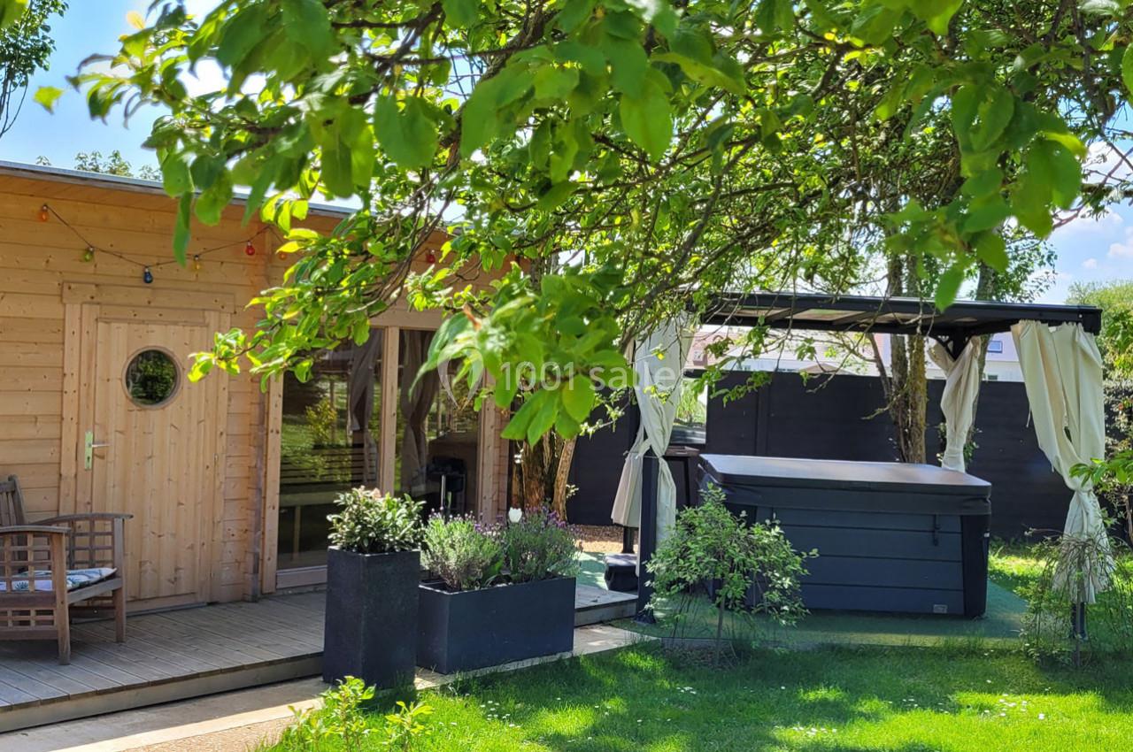 Location salle Bailleau-l'Évêque (Eure-et-Loir) - Le Domaine du Moulin 28 & SPA #11 Cabane en bois avec terrasse, entourée de verdure, un spa et un espace ombragé avec rideaux dans un jardin.