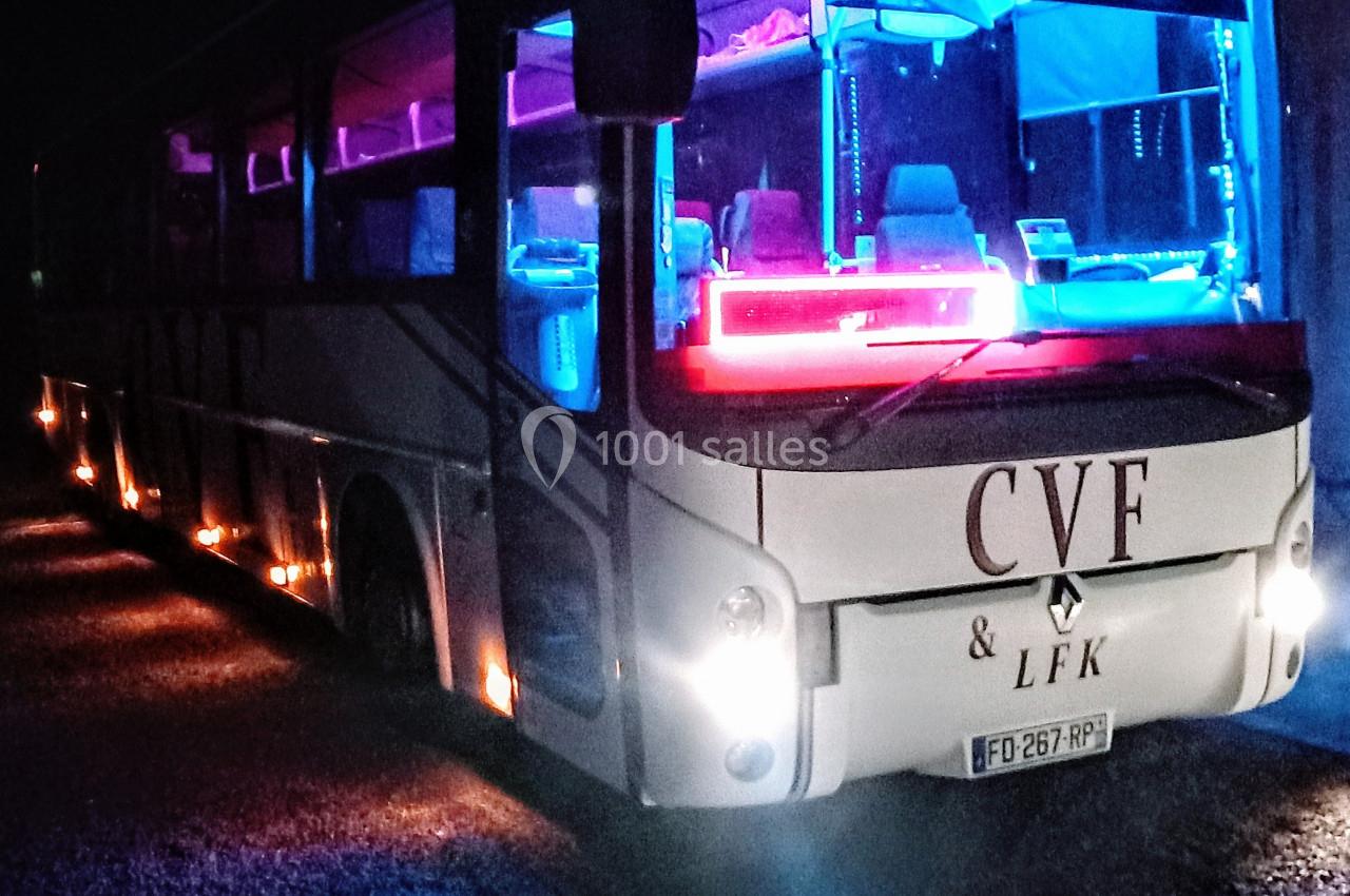 Autobus illuminé par des lumières colorées bleues et roses, stationné de nuit sur un sol gravillonné.