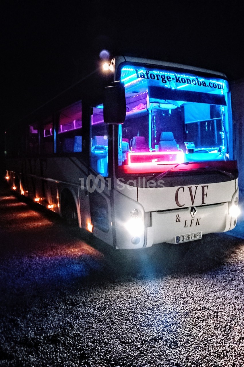 Autobus illuminé par des lumières colorées bleues et roses, stationné de nuit sur un sol gravillonné.