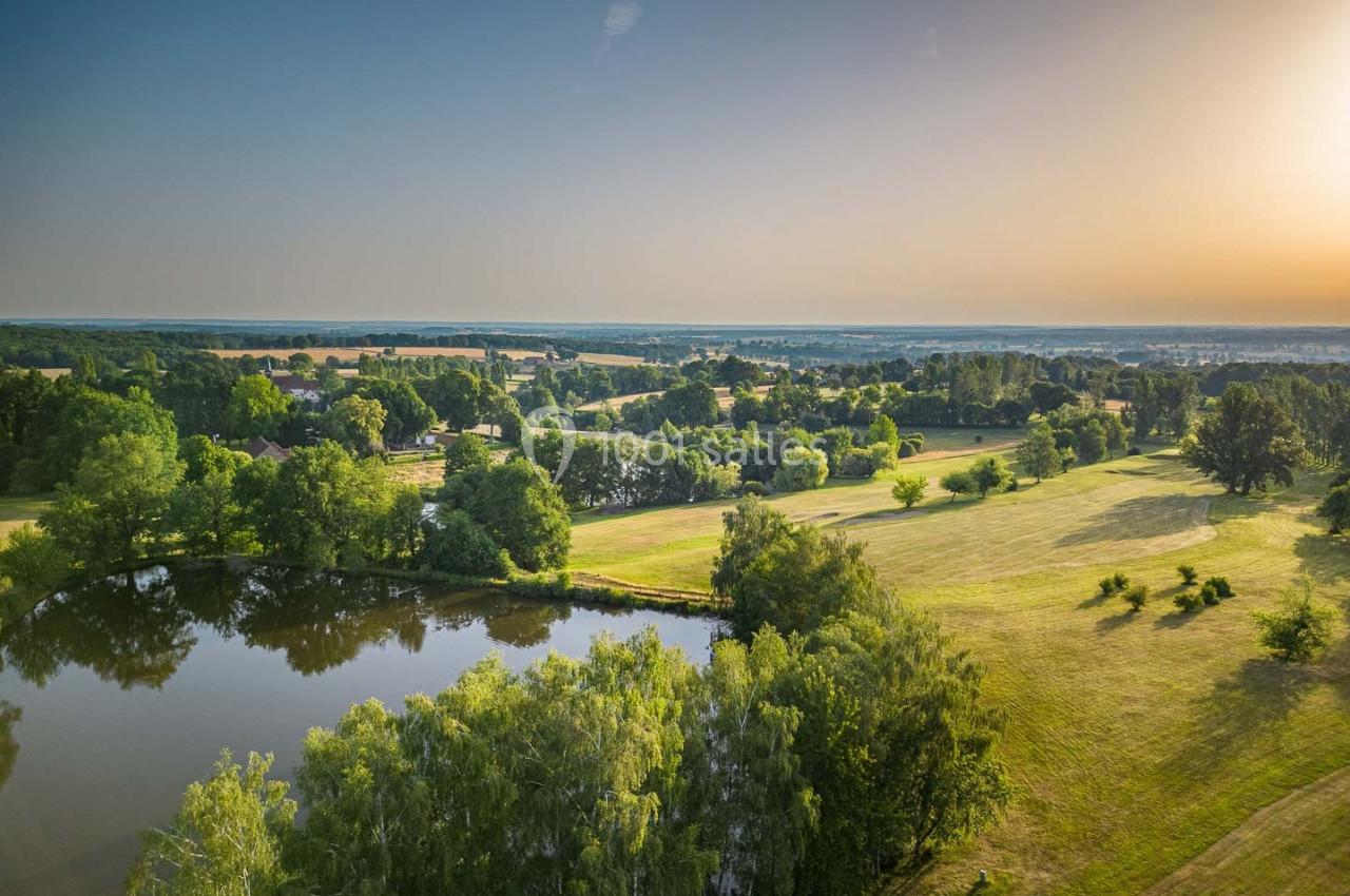 Vue aérienne d'un paysage rural avec un étang entouré d'arbres et des champs s'étendant à l'horizon sous un ciel dégagé.