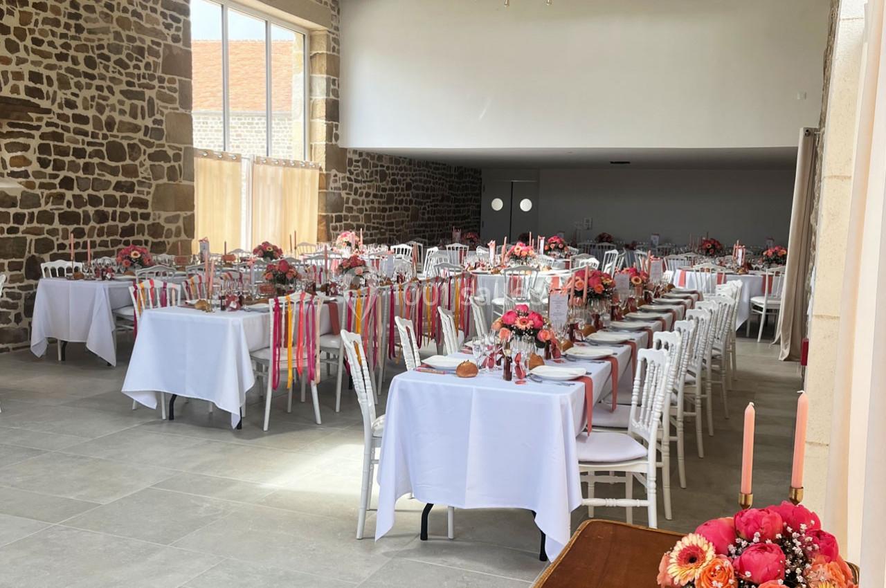 Salle de réception décorée pour un événement, avec des tables alignées, nappes blanches et arrangements floraux colorés.