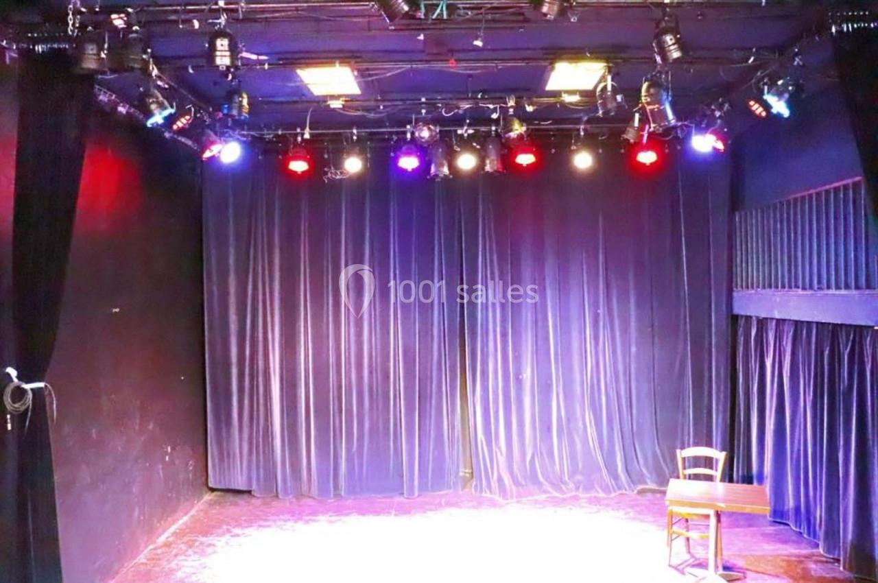 Scène de théâtre vide avec rideaux violets, éclairée par des projecteurs colorés, une chaise et une table en bois.