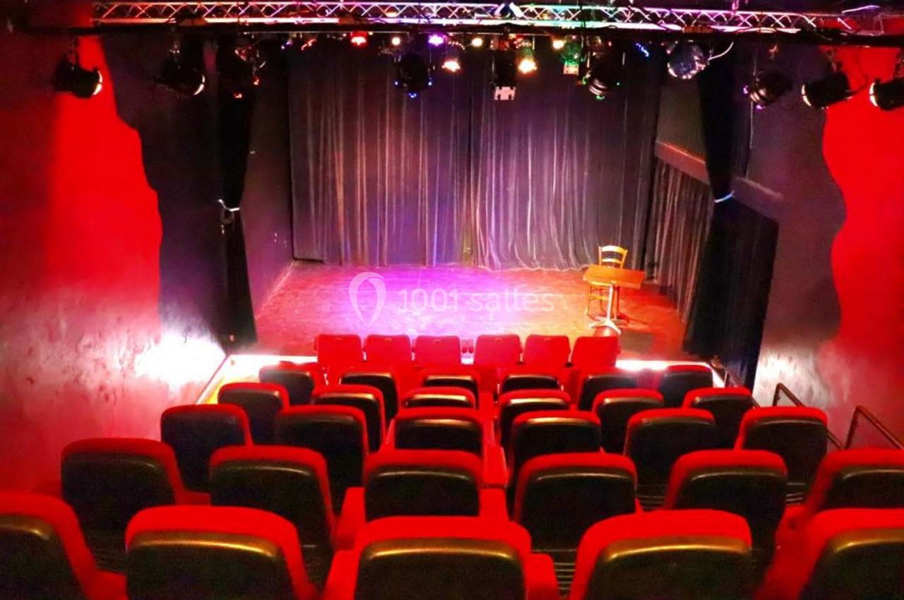 Salle de spectacle avec rangées de sièges rouges, scène éclairée et rideaux noirs en arrière-plan.