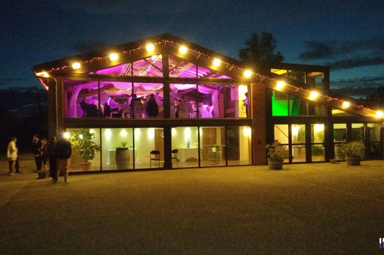 Bâtiment éclairé en soirée avec une salle vitrée illuminée de lumières colorées et des personnes visibles à l'intérieur.