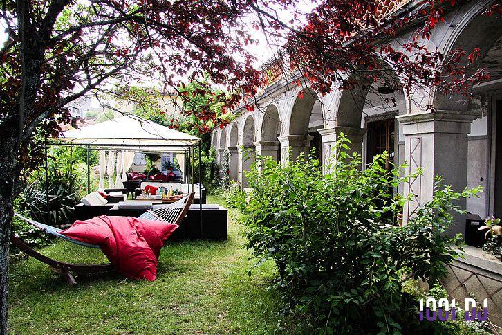 Jardin avec hamac, coussins rouges, mobilier de jardin sous une pergola et façade en arcades en arrière-plan.