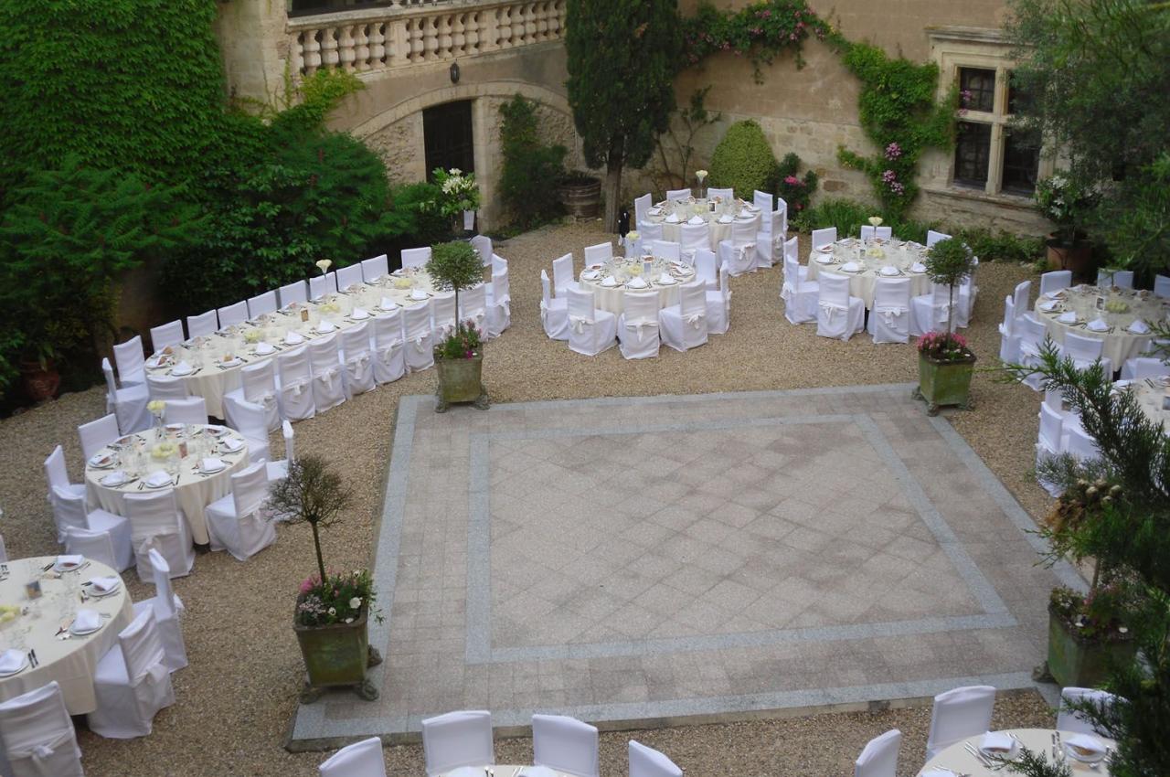 Tables rondes dressées avec nappes blanches dans une cour pavée entourée de murs en pierre et de végétation.