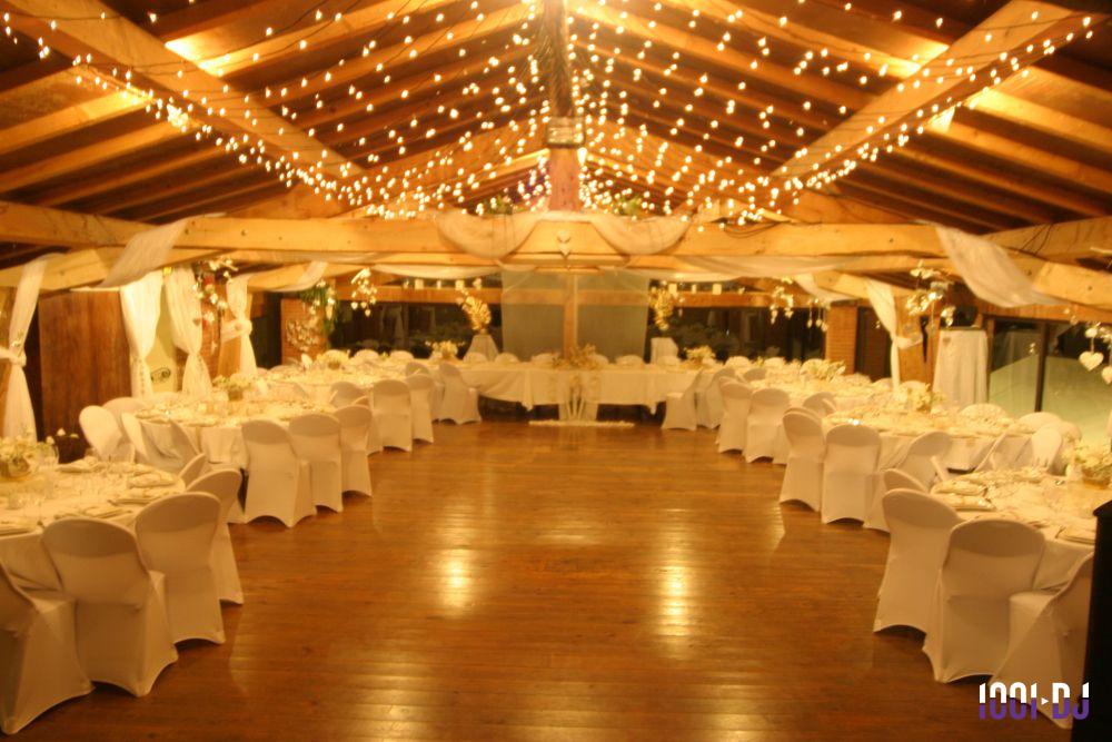Salle de réception décorée avec des guirlandes lumineuses, tables rondes et chaises blanches disposées en U.