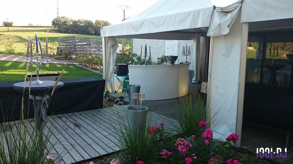Tente blanche avec bar arrondi sur terrasse en bois, entourée de fleurs et d'un jardin en arrière-plan.
