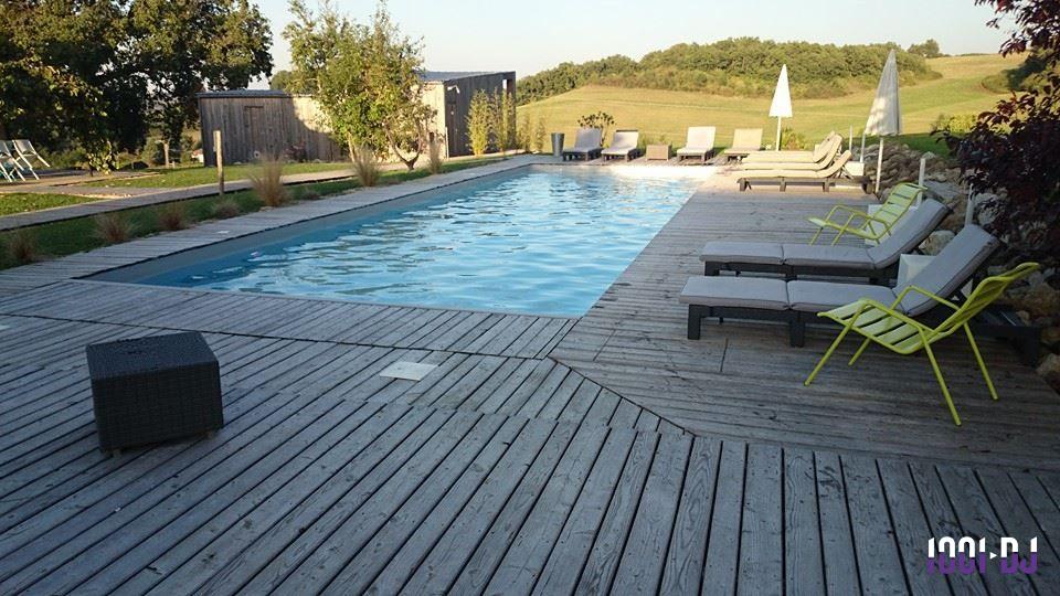 Piscine rectangulaire entourée de terrasses en bois avec chaises longues et paysage de campagne en arrière-plan.