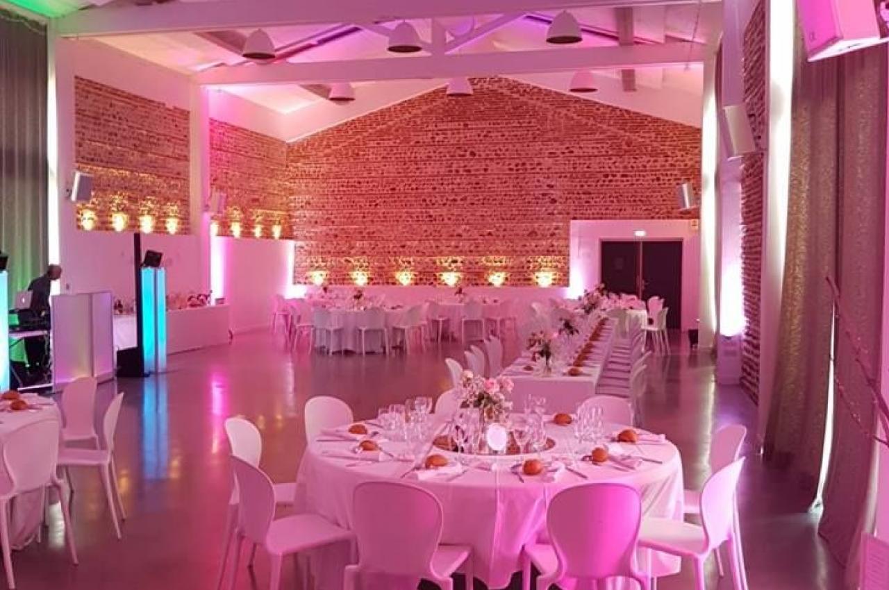 Salle de réception décorée avec des tables rondes dressées, éclairage rose et murs en briques apparentes.