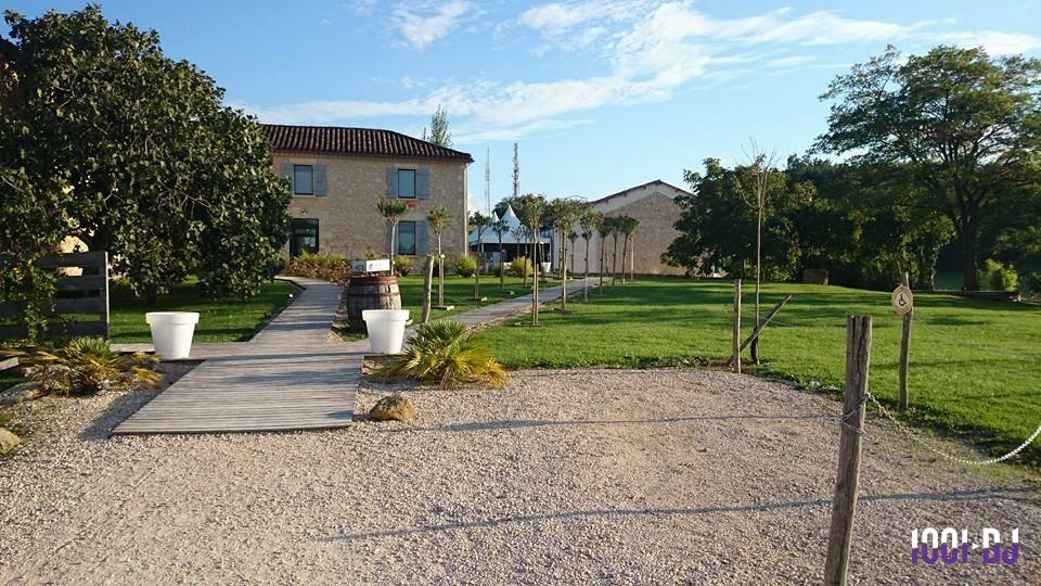 Allée en bois menant à une maison en pierre entourée de pelouse, d'arbres et de pots décoratifs blancs.