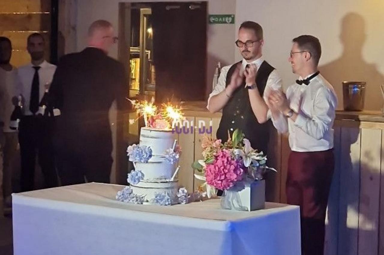 Deux hommes debout près d'un gâteau de mariage décoré, entouré de fleurs, avec des bougies allumées.