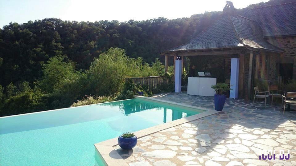 Piscine à débordement avec terrasse en pierre, vue sur une forêt et une petite structure en bois.