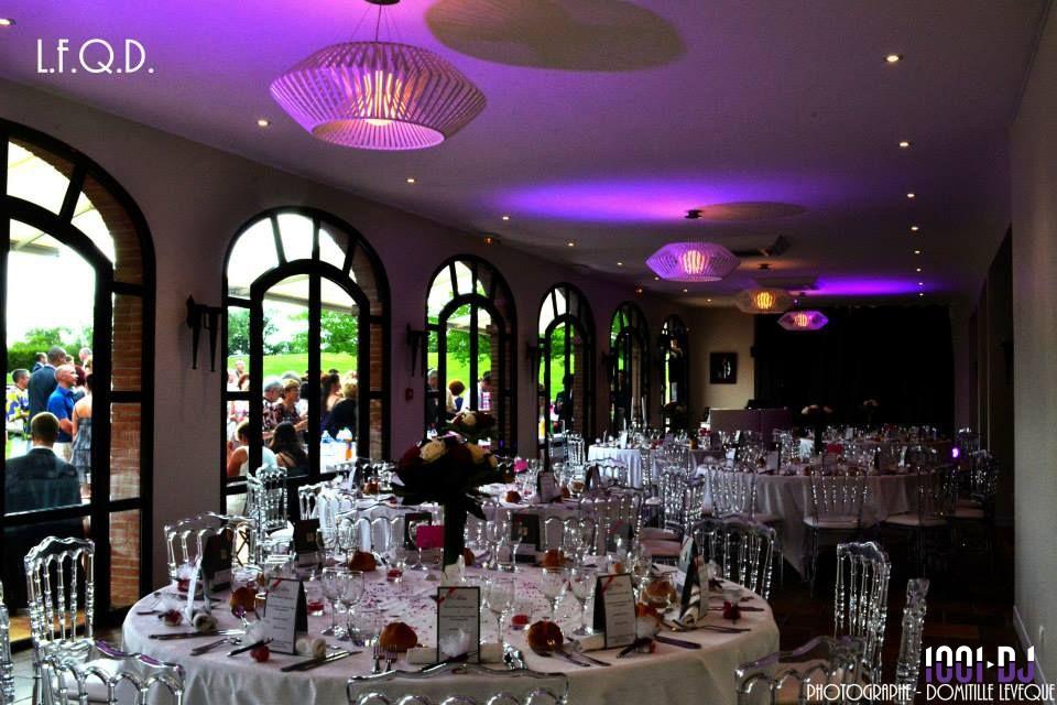 Salle de réception élégante avec tables dressées, chaises transparentes et éclairage violet, vue sur un jardin extérieur.