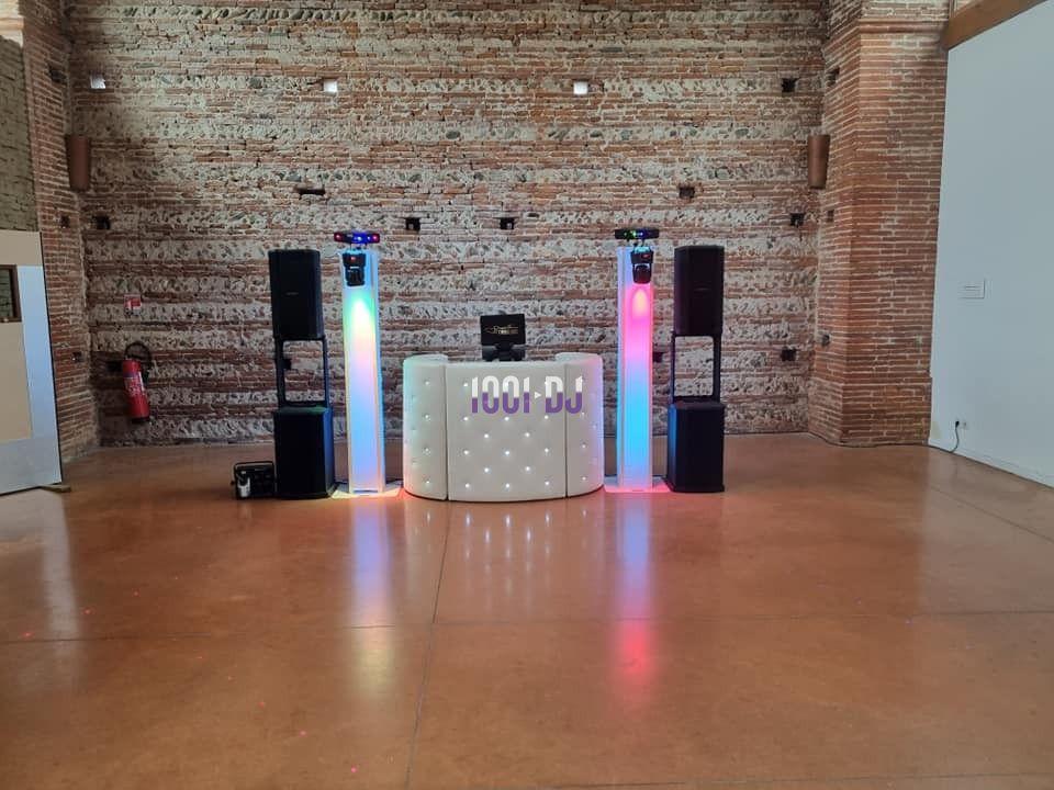 Console de DJ éclairée avec lumières colorées, entourée de deux colonnes lumineuses et d'enceintes, dans une salle en…