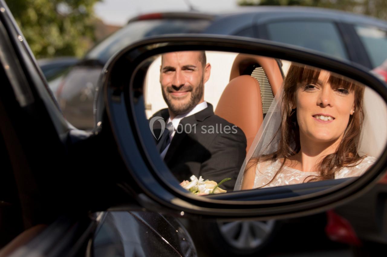 Vue dans un rétroviseur, un couple en tenue de mariage sourit à l'intérieur d'une voiture.
