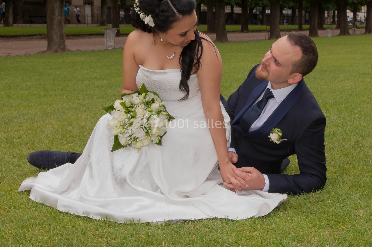 Un couple en tenue de mariage est assis sur une pelouse, se regardant avec complicité dans un parc arboré.
