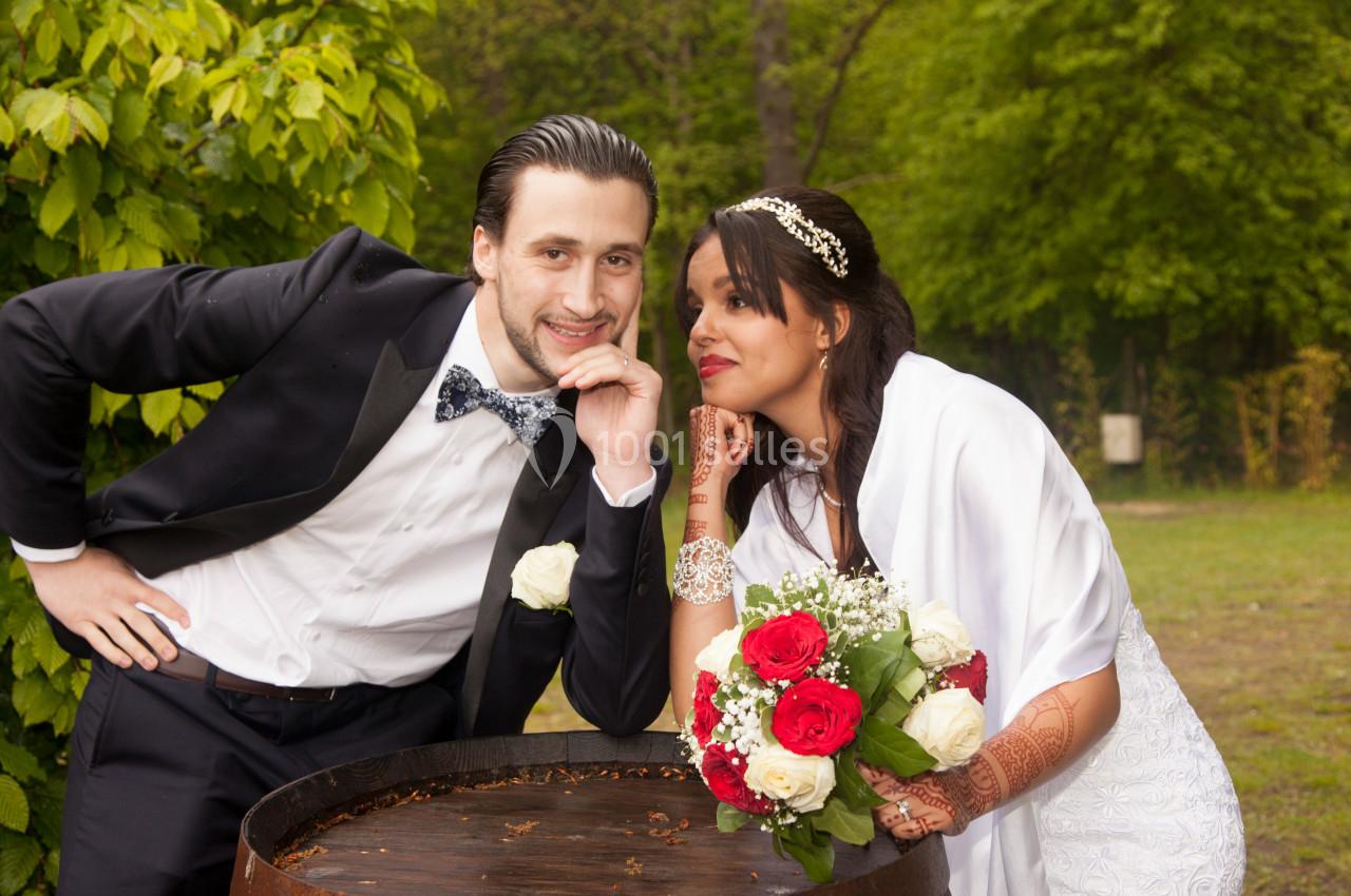 Un couple en tenue de mariage pose près d'un tonneau en bois dans un jardin verdoyant.