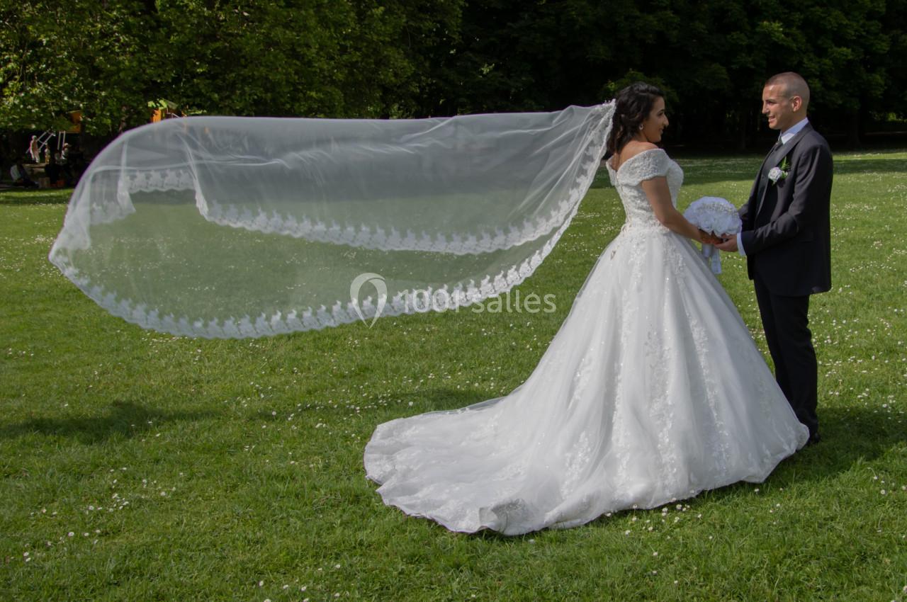 Une mariée en robe blanche avec un long voile se tient face à un homme en costume dans un parc verdoyant.