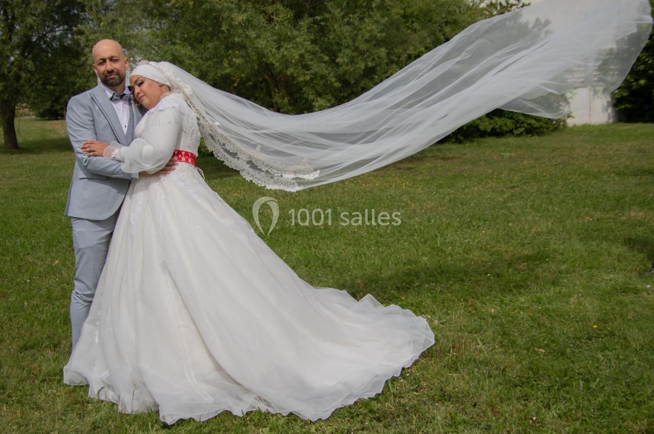Un couple en tenue de mariage pose dans un parc, la mariée portant un voile long flottant au vent.