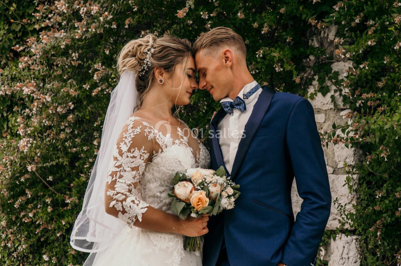 Un couple de mariés en tenue de cérémonie se tient face à face devant un mur fleuri.
