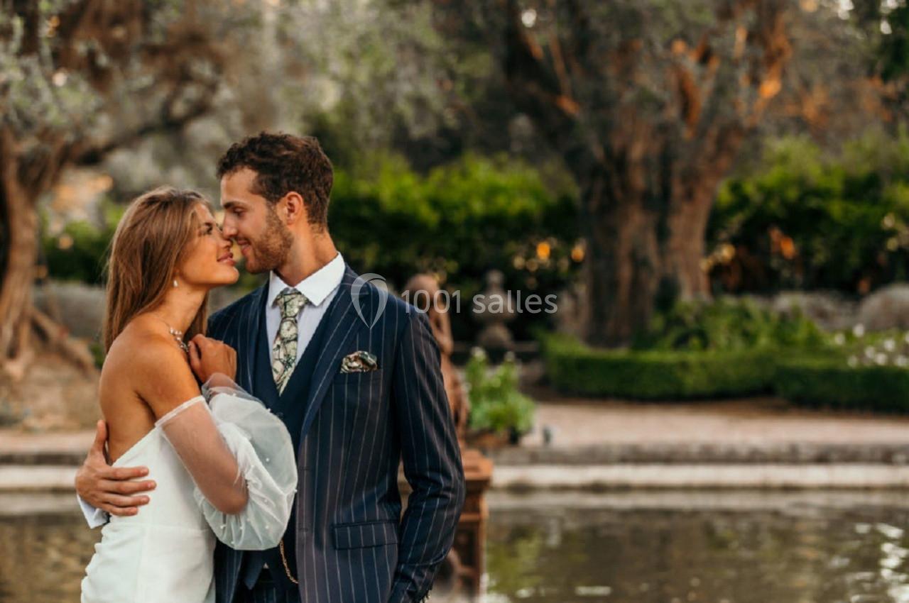 Un couple élégant s'embrasse tendrement devant un jardin verdoyant avec un bassin en arrière-plan.