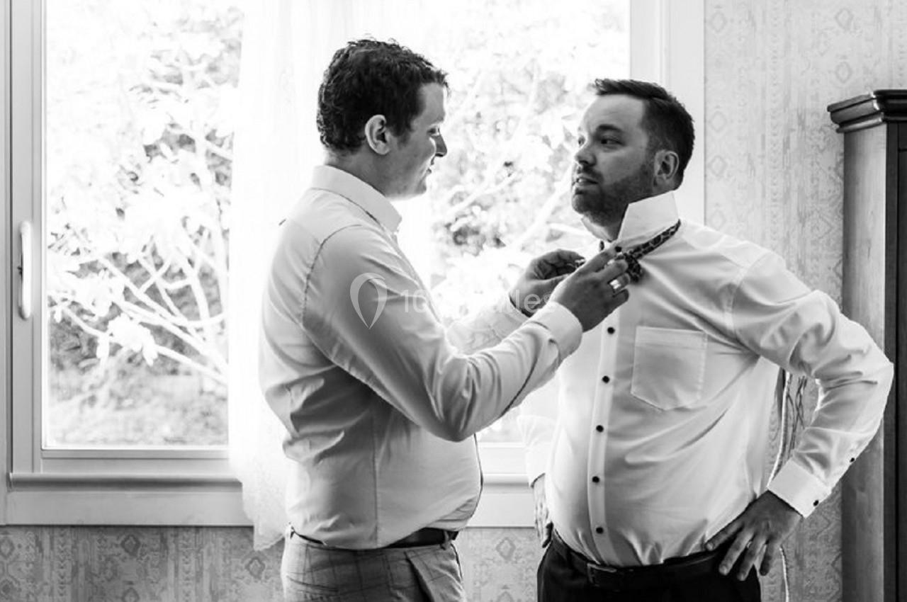 Un homme aide un autre homme à ajuster sa cravate dans une pièce lumineuse près d'une fenêtre.