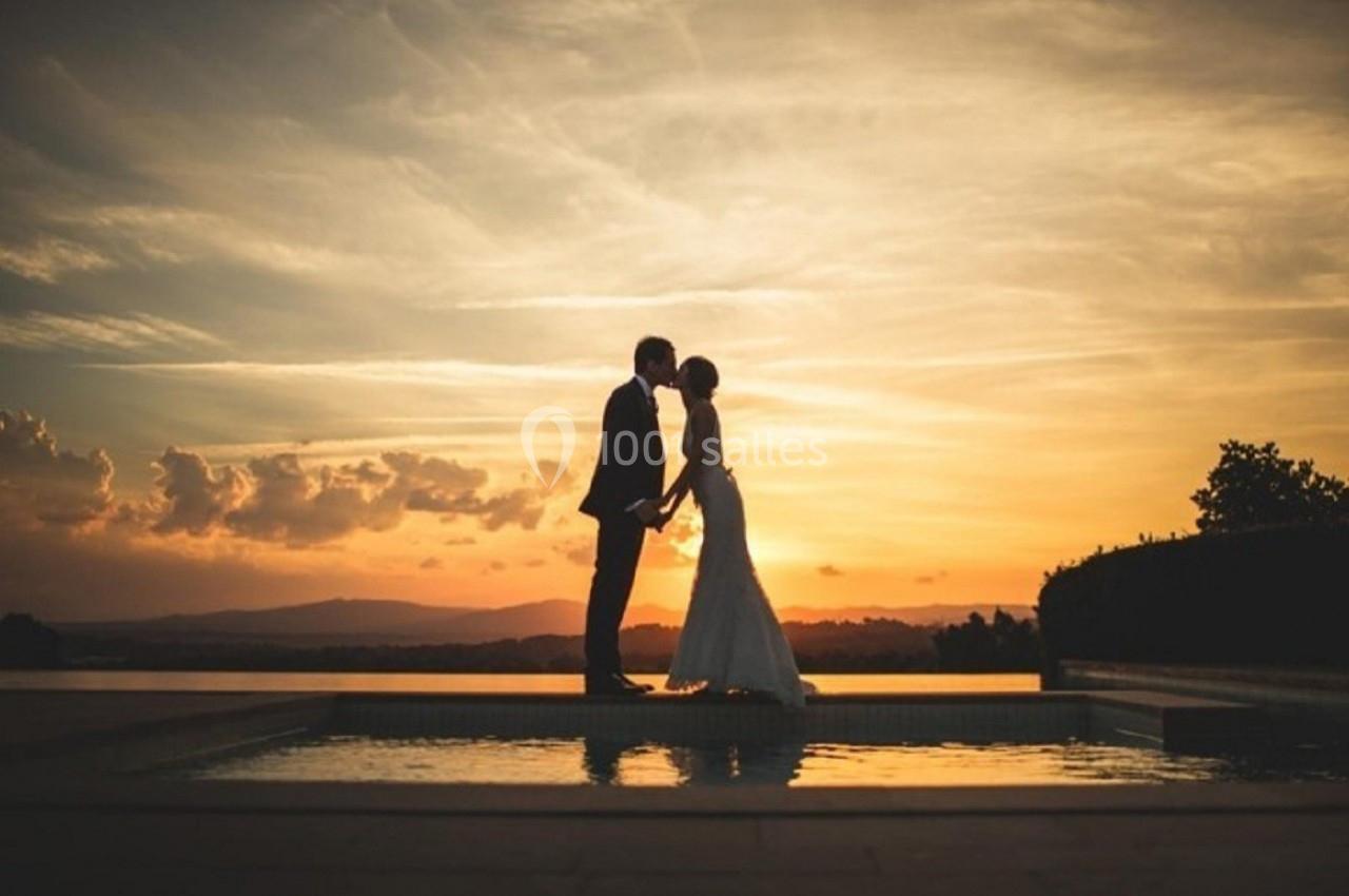 Un couple en tenue de mariage se tient près d'une piscine au coucher du soleil, avec un paysage en arrière-plan.
