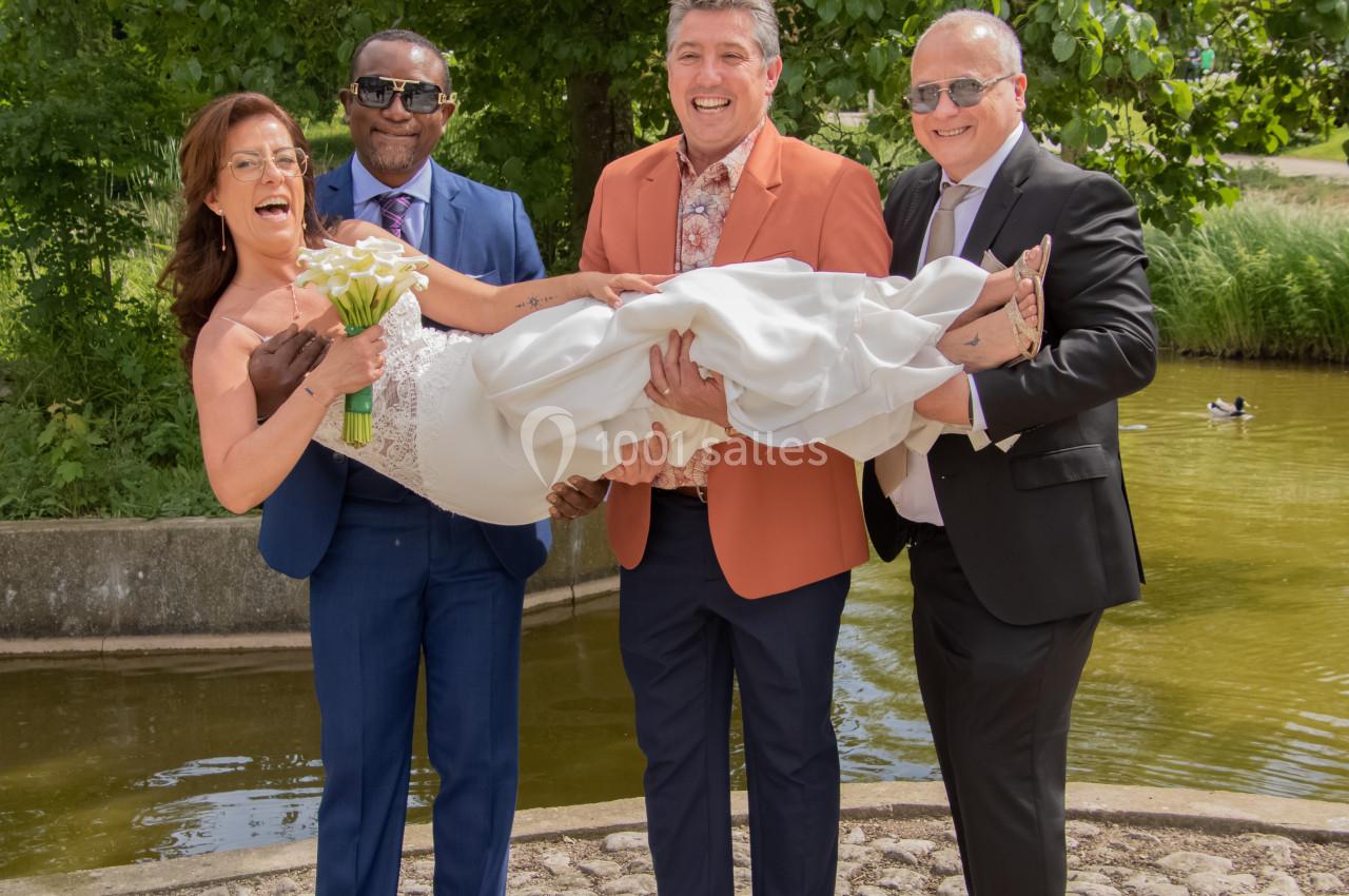 Une mariée en robe blanche est portée par trois hommes souriants devant un étang et des arbres.