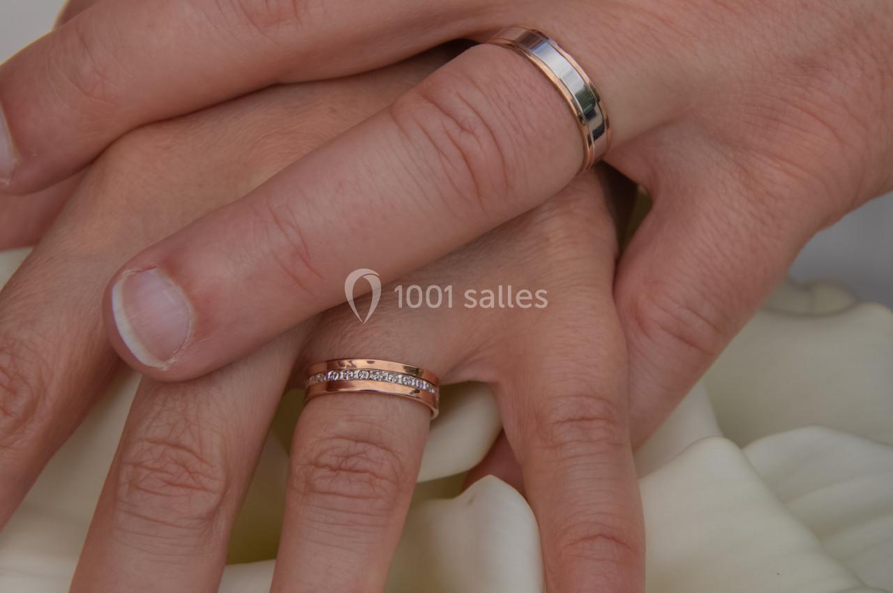 Deux mains portant des alliances en or rose, posées sur un bouquet de fleurs blanches.