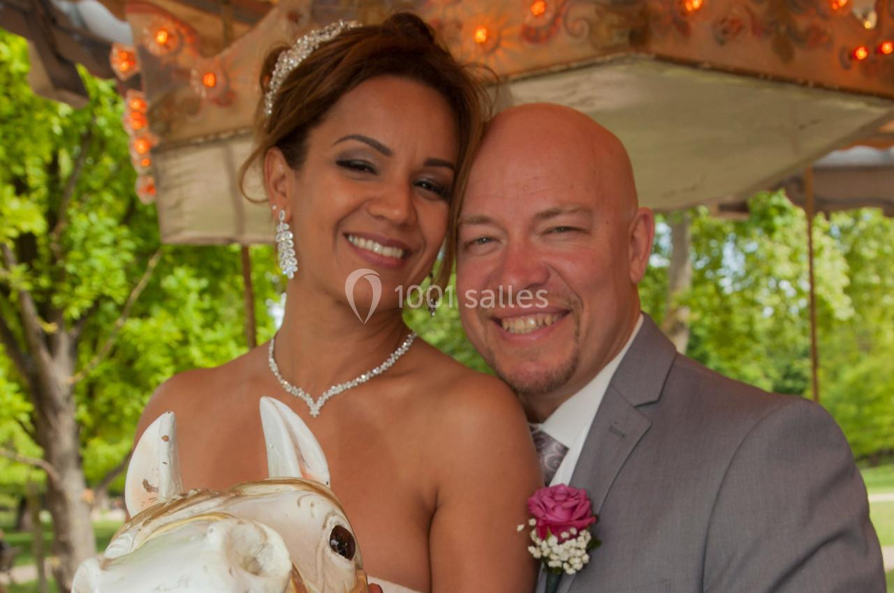 Un couple souriant pose sur un cheval de manège, avec un arrière-plan de verdure et de lumières décoratives.