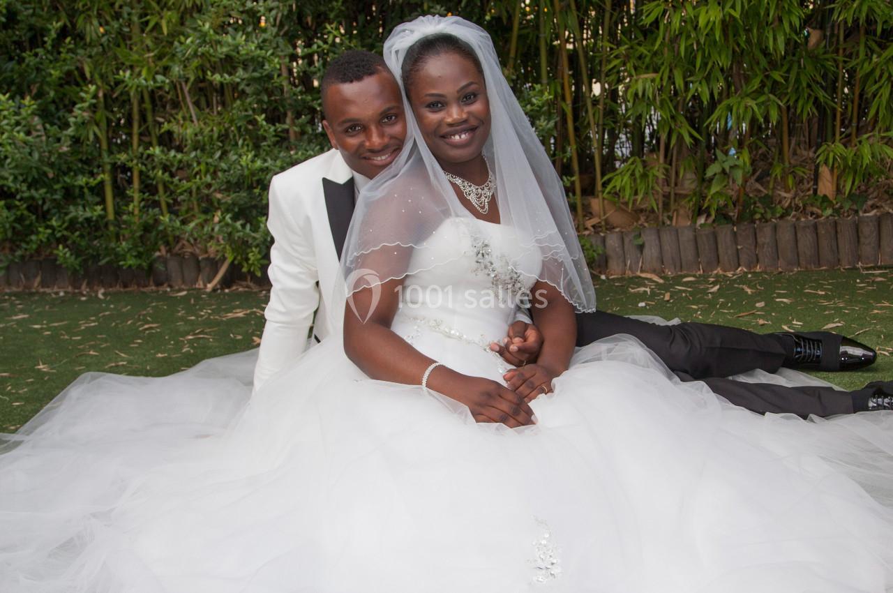 Un couple de mariés souriant, assis sur une pelouse, la mariée en robe blanche et le marié en costume clair.