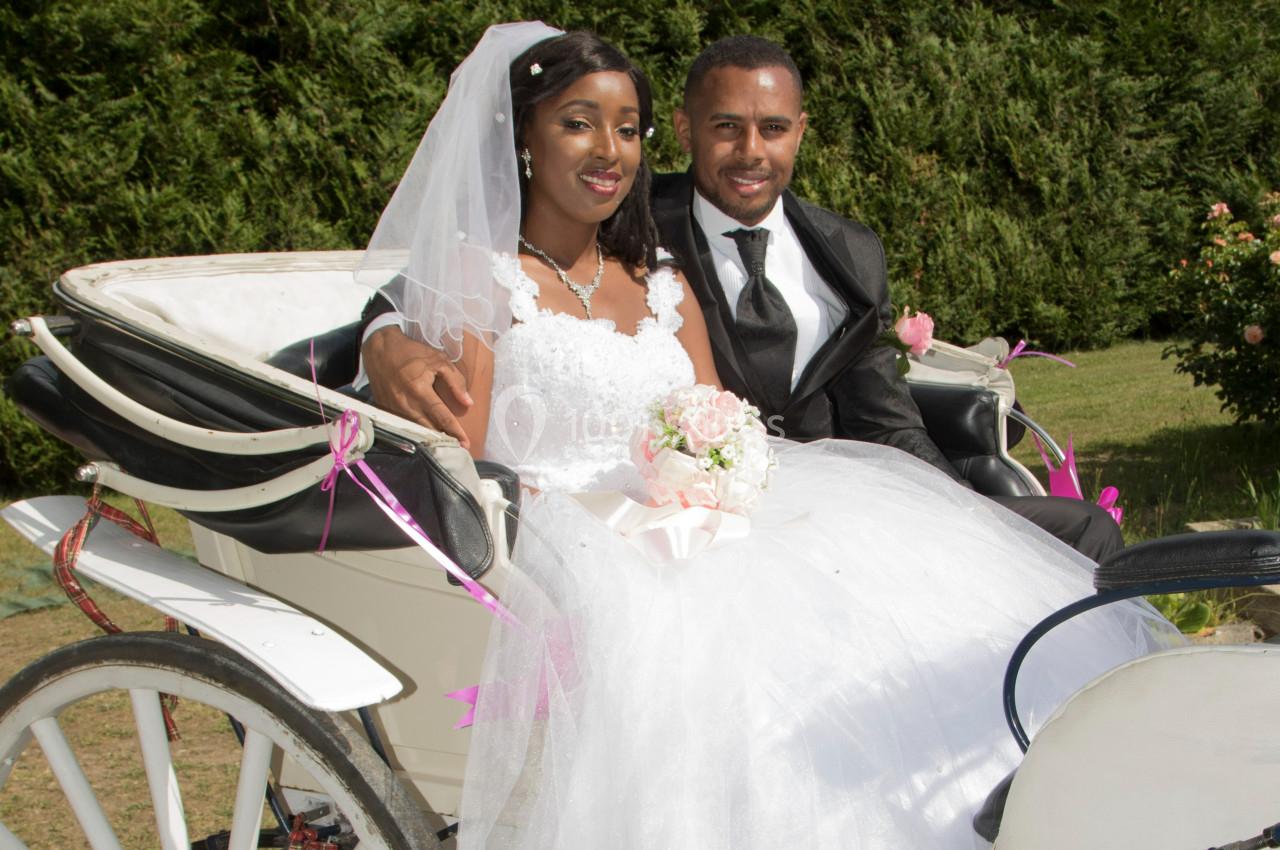 Un couple en tenue de mariage assis dans une calèche blanche décorée de rubans roses, en extérieur.