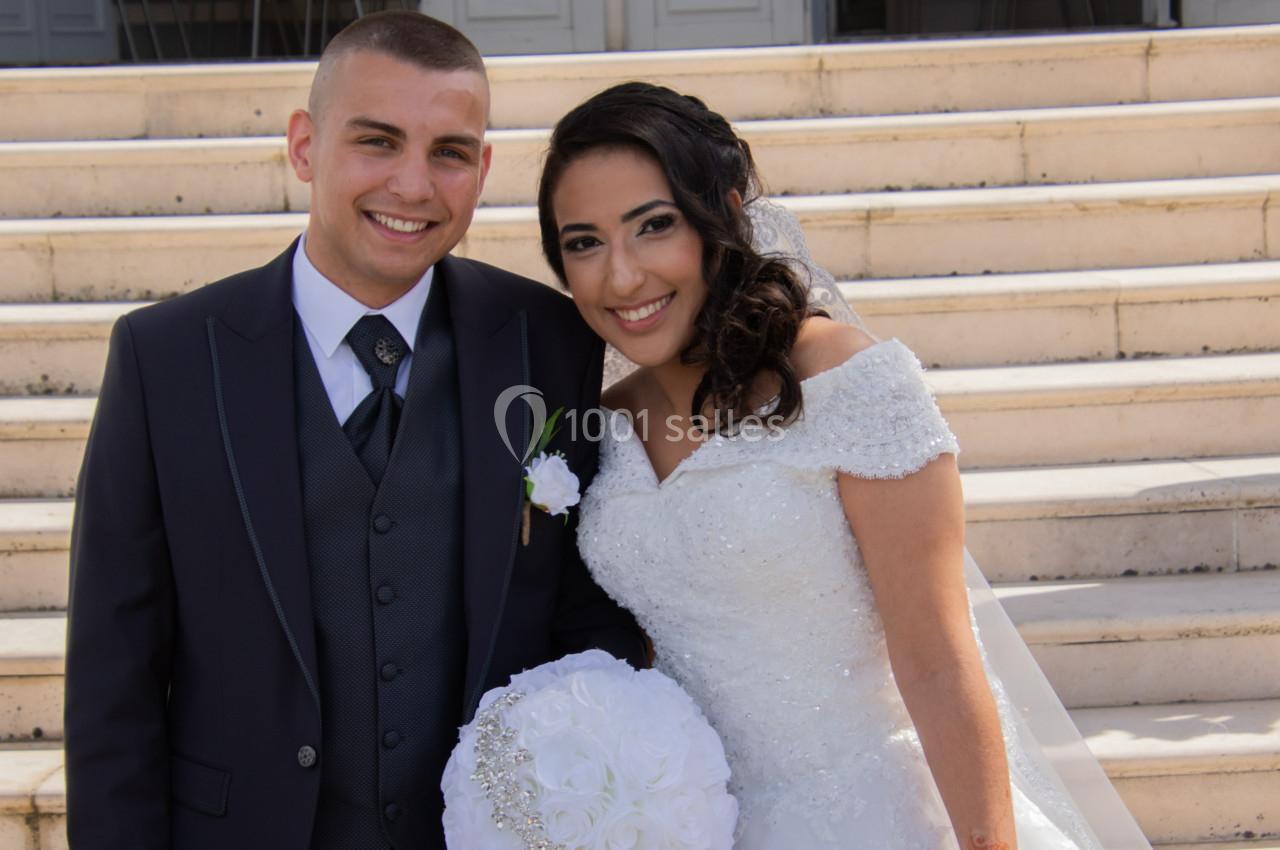 Un couple souriant en tenue de mariage pose devant des marches en pierre.