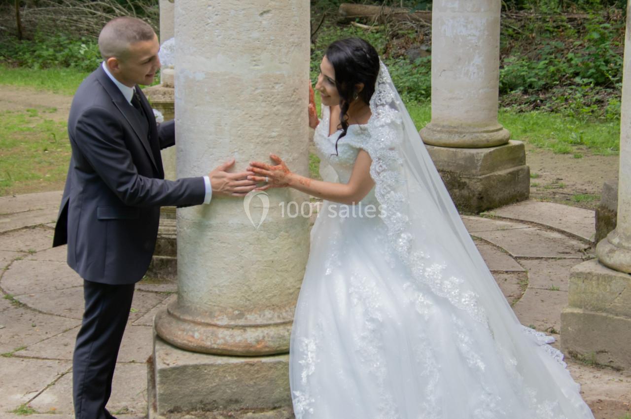 Un couple en tenue de mariage se tient près d'une colonne dans un jardin verdoyant.