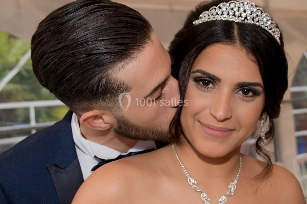 Un homme en costume embrasse tendrement une femme portant une robe et un diadème, dans un cadre festif.
