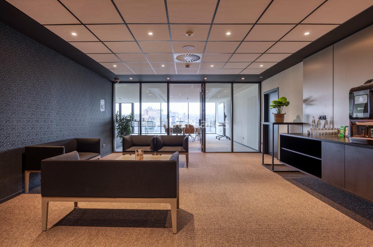 Salle d'attente moderne avec canapés noirs, table basse, plantes et vue sur une salle de réunion vitrée.