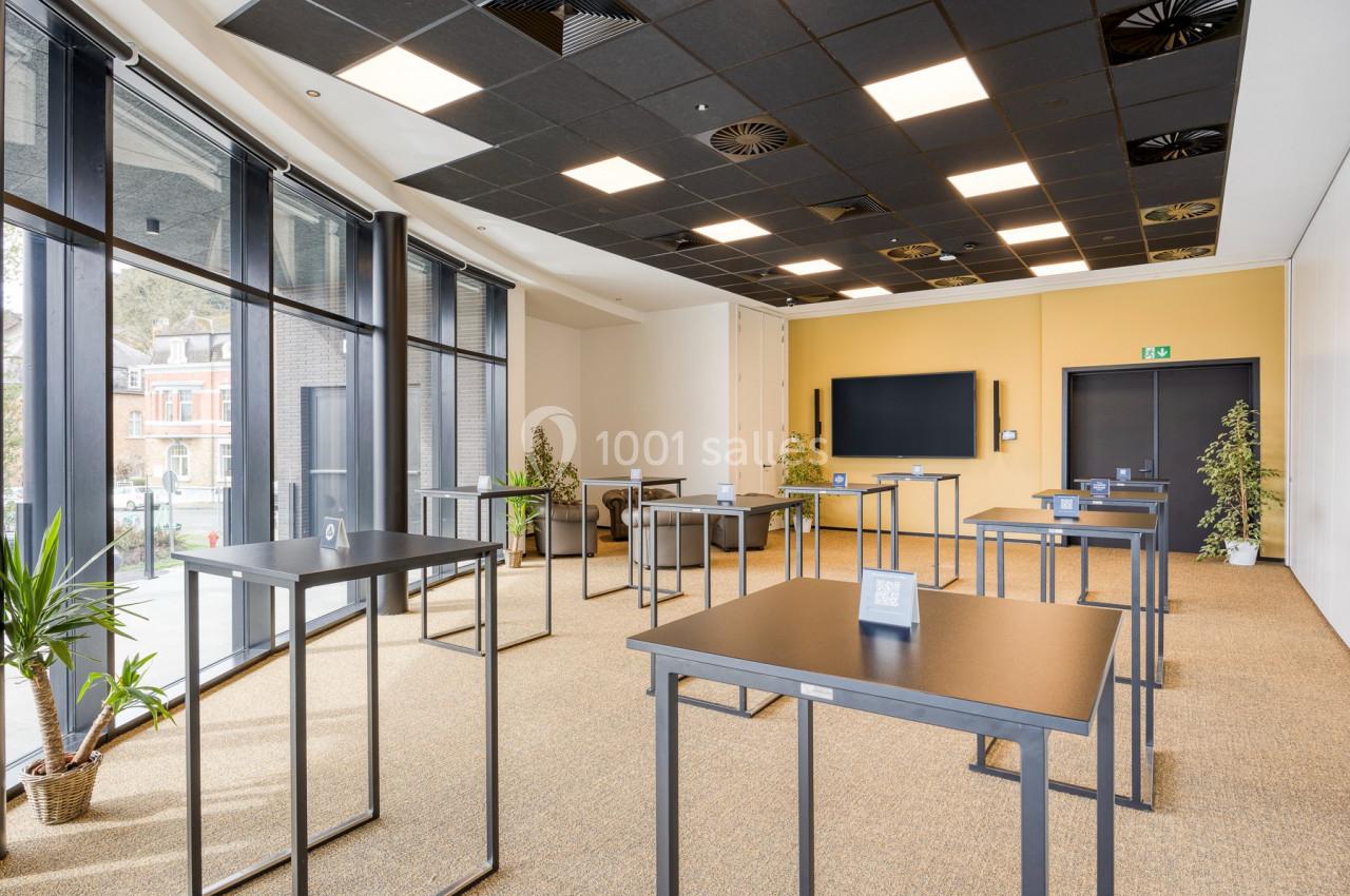 Salle de réunion lumineuse avec tables hautes, grandes fenêtres, plantes et écran mural.