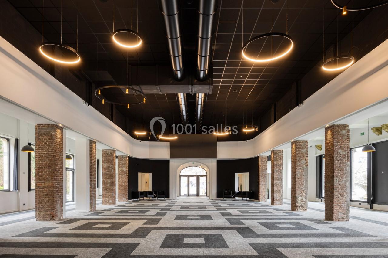 Grande salle moderne avec plafond noir, colonnes en briques, éclairage circulaire suspendu et sol à motifs géométriques.