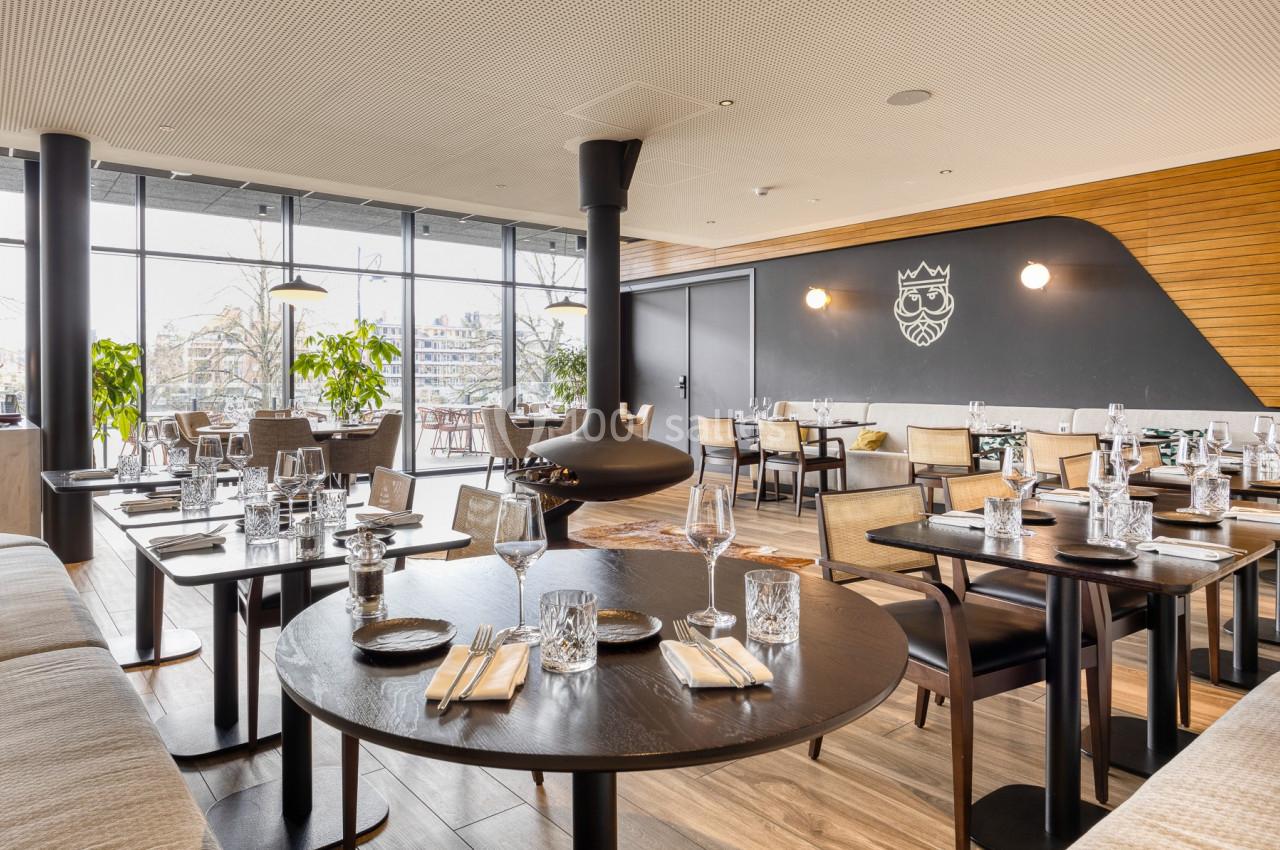 Salle de restaurant moderne avec tables dressées, grandes baies vitrées et cheminée suspendue au centre.