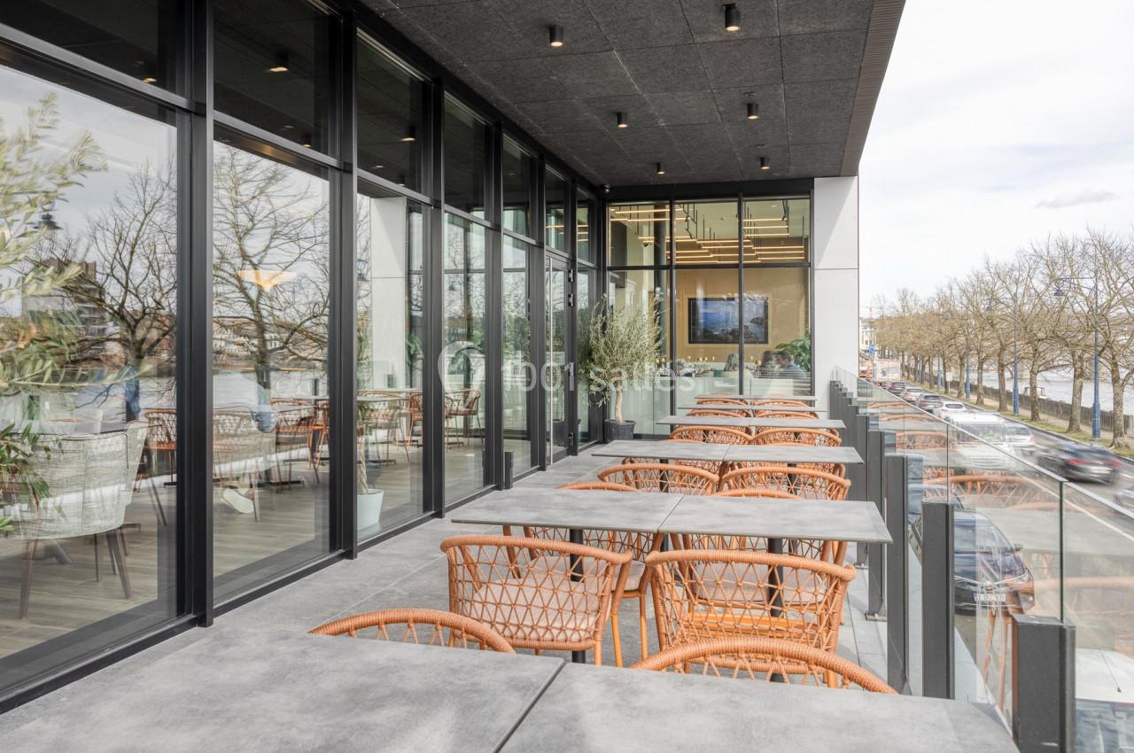 Terrasse moderne avec tables en béton et chaises en rotin, bordée de baies vitrées et offrant une vue sur des arbres et une…
