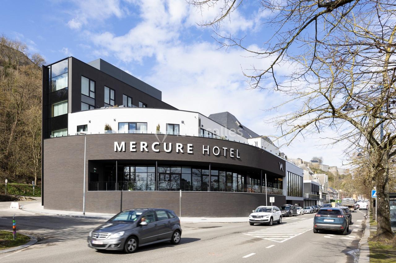 Façade moderne d'un hôtel Mercure avec des voitures circulant devant, entouré d'arbres et d'un ciel partiellement nuageux.