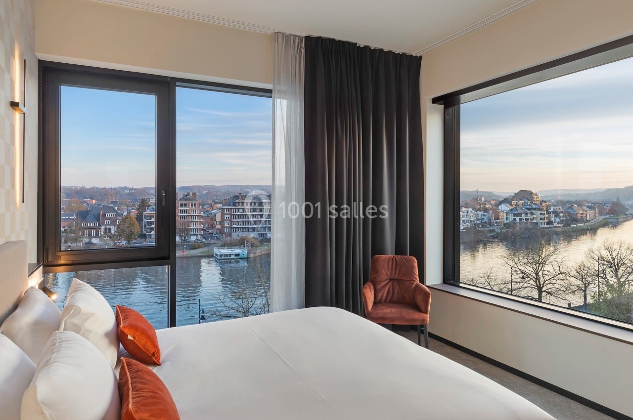Chambre lumineuse avec lit double, fauteuil et grandes fenêtres offrant une vue sur une rivière et des bâtiments…
