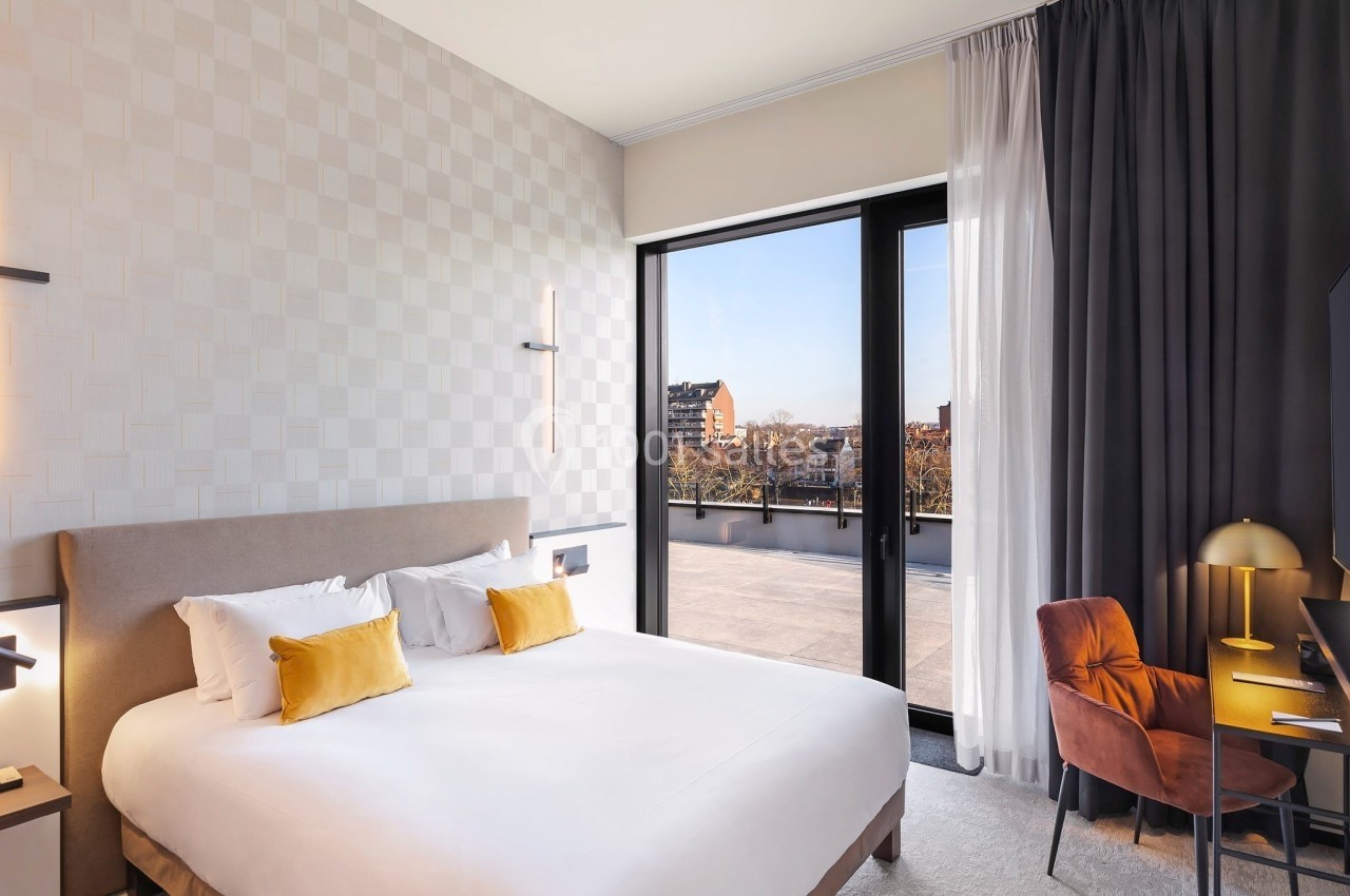 Chambre lumineuse avec lit double, bureau, chaise et grande baie vitrée donnant sur une terrasse et vue urbaine.